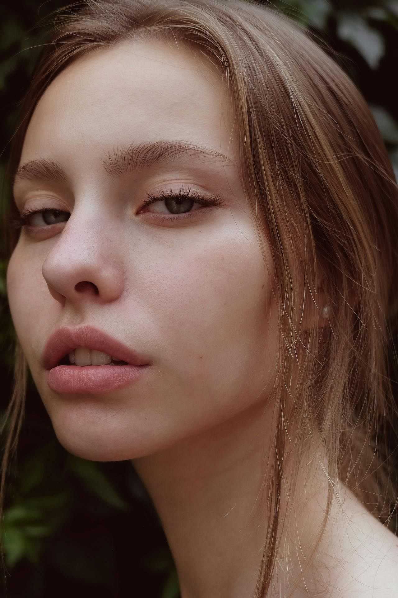 Close-up of a woman with light brown hair gazing confidently, green foliage in the background.